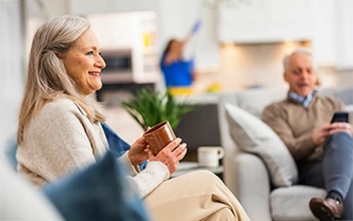 Women sitting in her living room drinking coffee and smiling while a maid cleans in the kitchen