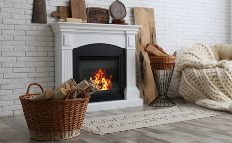 A fire crackles in the fireplace with a basket of wood and chair with woven blanket on each side of the fireplace.